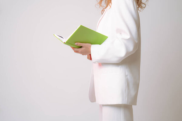 Side view photo of a female in white suit with green notebook in her hands, isolated over grey background