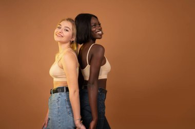 Two cheerful international girls friends in casual clothes standing back to back and smiling, isolated over dark orange background