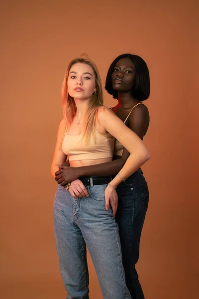 Dos chicas internacionales serias en ropa casual, chica africana abrazando a la rubia europea, ambas mirando a la cámara, aisladas sobre fondo naranja —  Fotos de Stock