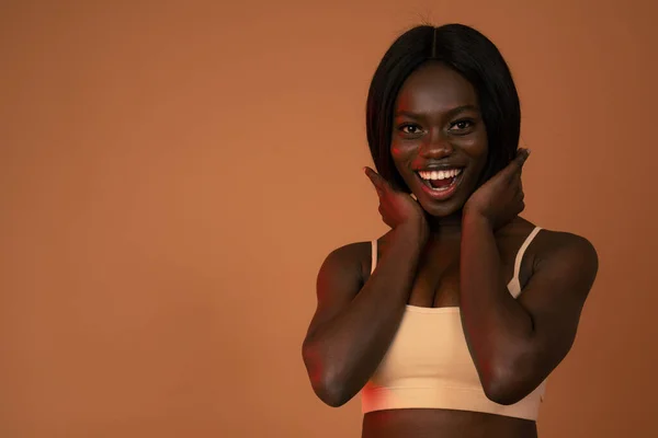 Waist up portrait of a happy attractive african girl in beige bra who holds her hands on her neck, smiling and looking at the camera isolated over orange background — 图库照片
