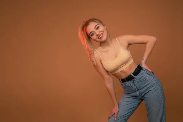 Привлекательная блондинка в бежевом топе и синих джинсах опиралась на ногу, улыбаясь и глядя на камеру, изолированную на оранжевом фоне — стоковое фото