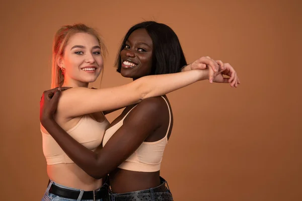 Beauty portrait of young happy optimistic cheery beautiful blonde european and brunette african women smiling and looking at the camera isolated over orange wall background — Stockfoto