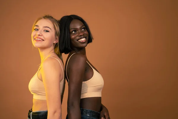 Filles internationales agréables en soutien-gorge beige, filles attrayantes africaines et européennes debout dos à dos et souriant, isolé sur fond orange avec copyspace — Photo