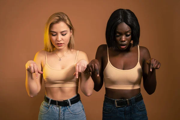 Shocked young international girls in casual clothes posing isolated on yellow orange background in studio. People lifestyle concept. Mock up copy space. Pointing index fingers and looking down — Stockfoto