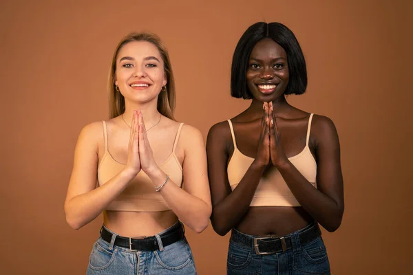 Ung glad blond europeisk kvinna och afrikansk leende flicka praktiserar yoga, håller händerna i namaste och håller ögonen stängda. Kvinnor mediterar inomhus, ber för fred och kärlek, har lugn — Stockfoto
