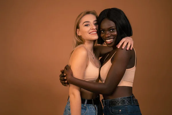 Feliz internacional sonriente atractiva pareja de lesbianas en sujetador beige, chicas europeas y africanas abrazando y mirando a la cámara aislada sobre fondo naranja oscuro —  Fotos de Stock