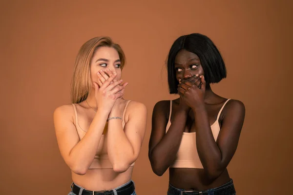 Waist up photo of two international girls in beige bra closing their mouths with their hands and looking at each other