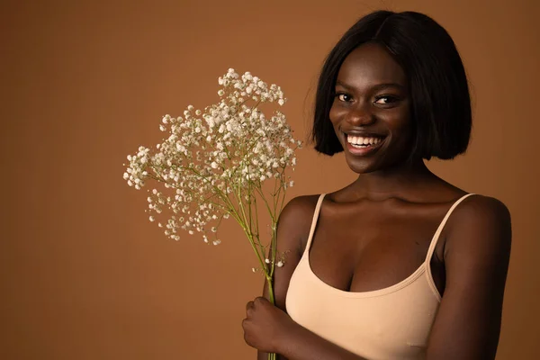 Photo of pleasant looking dark skinned woman with cheerful facial expression, satisfied with something, carries white spring flowers, wears beige bra, isolated over blue background. — 图库照片