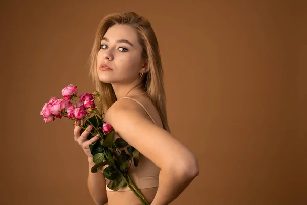 Gorgeous blonde woman with healthy fresh skin holds pink flowers and looking at the camera over her shoulder, isolated over orange background — Stockfoto