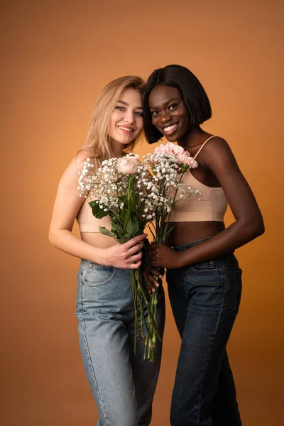 Happy smiling girls in blue jeans and beige top looking at the camera, european and african girls holding flowers — Stockfoto