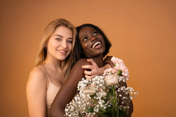 European blonde girl hugging smiling african smiling woman with flowers isolated on orange background — Stockfoto