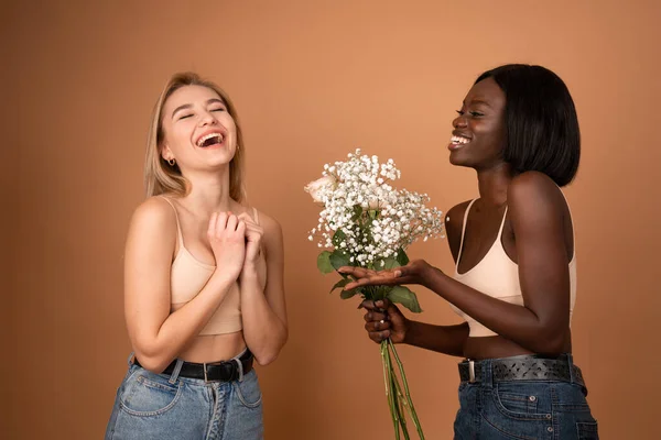 Overjoyed lovely young european lady receives congratulation and flowers on birthday, african smiling woman stretches hands and gives spring white flowers, isolated over orange background. — Stockfoto