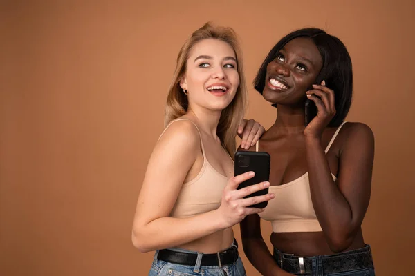 Two happy attractive international girls wearing beige bra standing isolated over dark orange background, using mobile phone — Stockfoto