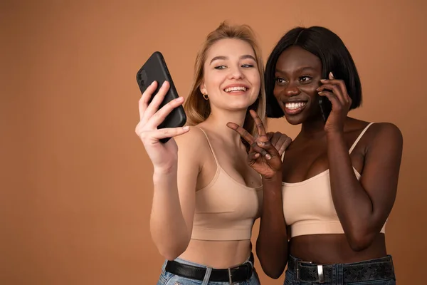 Портрет счастливых двух улыбающихся международных девушек, делающих селфи на смартфоне и показывающих два пальца на оранжевом фоне — стоковое фото