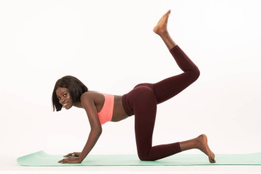 Yandan bakıldığında yoga minderinde yoga yapan, kameraya bakan spor giyimli zayıf Afrikalı bir kızın fotoğrafı.
