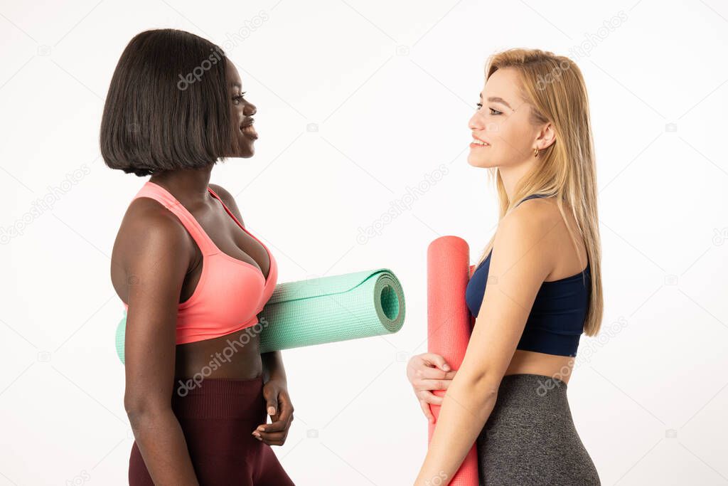 Sporty international girls holding yoga mats and looking at each other wearing workout clothing over vibrant whire background. Fashion, sport and healthy lifestyle concept