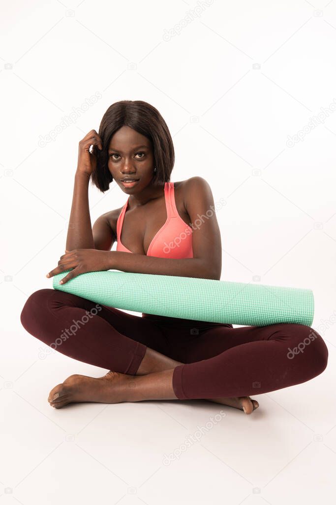 Cute fit young dark skinned woman in trendy sports clothes sitting with mat with legs folded. Healthy active black girl relaxing in padmasana after yoga practice, looking at camera