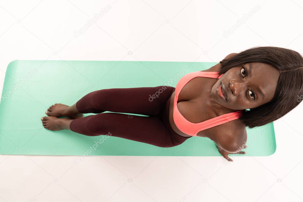 Top view photo of young african girl in stylish sportswear who sitting om the yoga mat and looking at the camera