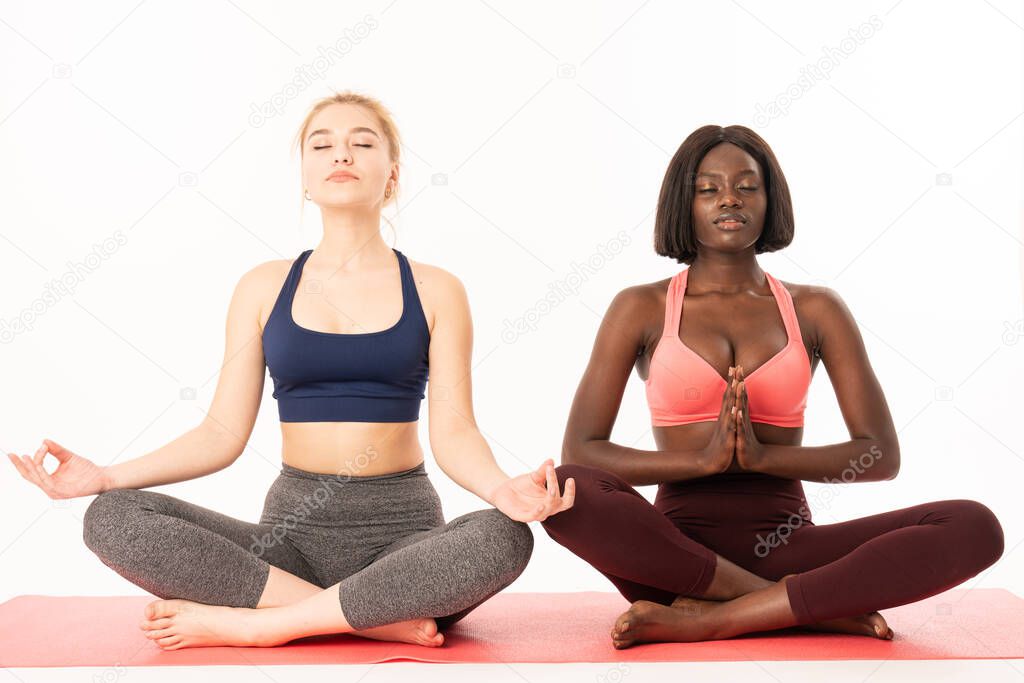 International girls in sportswear sitting in lotus position, african and european females meditating with closed eyes while sitting on a yoga mat