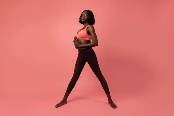 Full längd foto av en sportig afrikansk flicka i sportkläder står med breda ben, hålla händerna tillsammans och tittar seriöst på kameran, isolerade över rosa bakgrund — Stockfoto