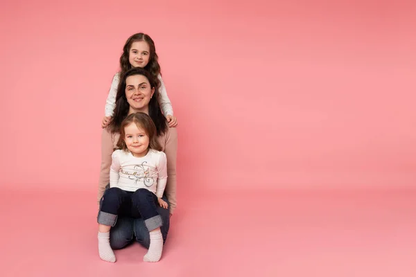Vacker ung mor med sina två döttrar sittande på marken en efter en. Titta rakt på kameran. Isolerad på rosa bakgrund — Stockfoto