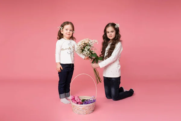 Två små systrar i casual kläder med blommor i håret står nära en korg med våren blommor, ger glad flicka blommor till sin söta syster — Stockfoto