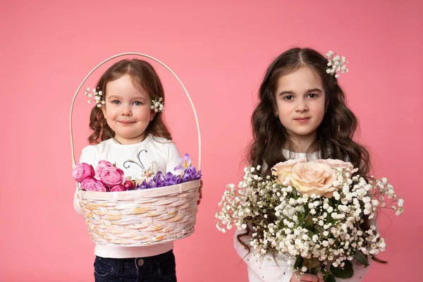 Två vackra små systrar med blommor i lockigt hår håller bukett och korg i händerna och tittar på kameran, vår skönhet porträtt — Stockfoto