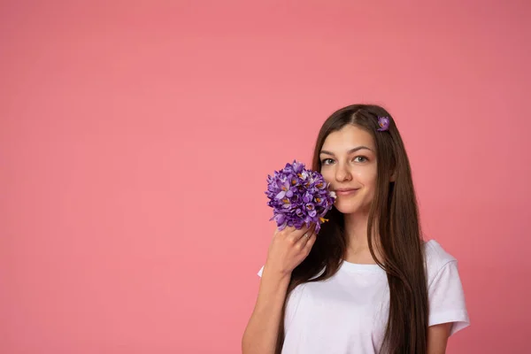 Mulher morena alegre em camiseta branca segurando flores de gotas de neve de primavera e olhando para a câmera, isolada sobre fundo rosa — Fotografia de Stock