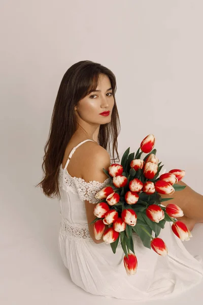 Confident brunette female with red lips dressed in white dress, holding tulips in her hands and looking at the camera over her shoulder — Stock Photo, Image