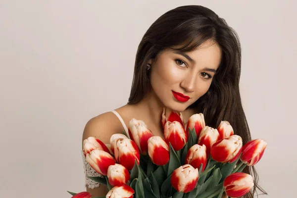 Közelkép tavaszi portré egy gyönyörű barna nő piros ajkak tartja piros tulipán és nézi a kamerába — Stock Fotó