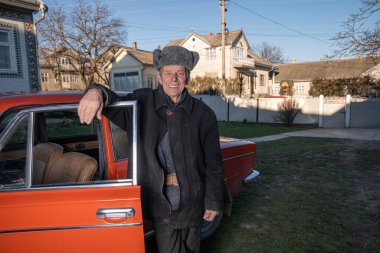 Siyah ceketli yaşlı bir adam ve gri şapkalı kırmızı retro arabasına yaslanmış, gülümsüyor ve kameraya bakıyordu