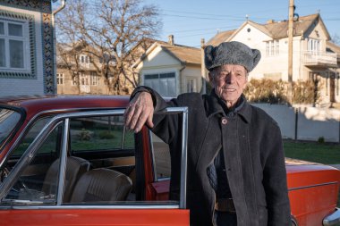 Gri retro şapkalı yaşlı adam kırmızı eski arabasının yanında bahçesinde duruyor.