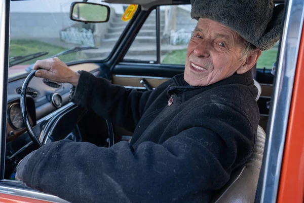 Hombre muy viejo y confiado en chaqueta negra y sombrero gris sentado en su coche retro y mirando a la cámara — Foto de Stock