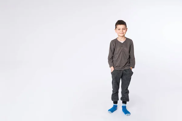 Liten attraktiv pojke i casual kläder håller händerna i fickor och tittar på kameran, isolerad på vit bakgrund — Stockfoto