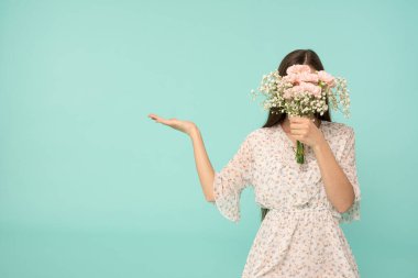 Parlak elbiseli genç bayan yüzünü bahar çiçekleri buketine gizler ve ürününüzü mavi arka planda izole bir şekilde açık avuçta gösterir.