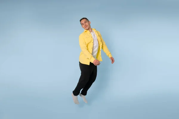 Грайливий молодий чоловік у жовтій сорочці стрибає і дивиться на камеру, ізольований на синьому фоні — стокове фото