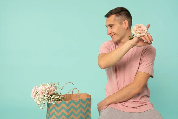 Atractivo joven en camiseta rosa con rosa en la mano apuntando a la esquina superior derecha, sentado en el suelo cerca de la bolsa de compras con flores y mirando al lado izquierdo — Foto de Stock