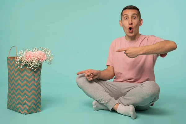 Hombre guapo sorprendido en camiseta rosa sentado en el suelo con la boca abierta, apuntando a la bolsa de compras con flores y mirando a la cámara — Foto de Stock
