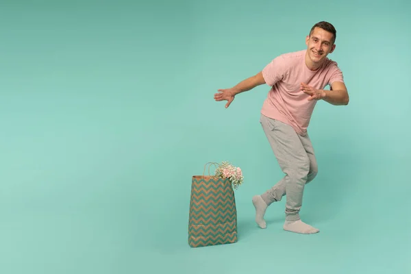 Hombre atractivo feliz en camiseta rosa y pantalones grises bailando cerca de la bolsa de compras con flores, aislado sobre fondo azul — Foto de Stock