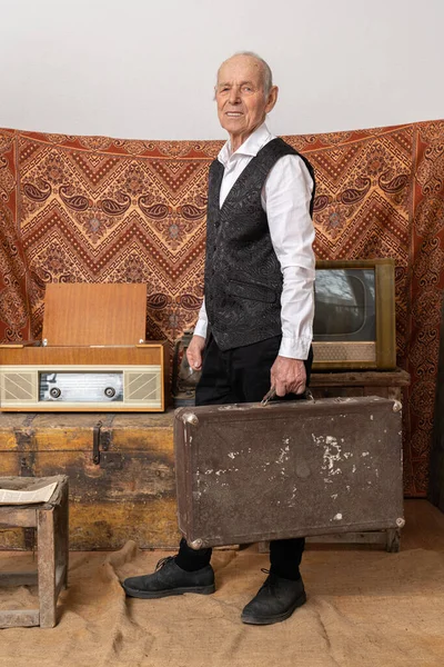 Старик в белой рубашке держит старый классический багаж путешествия, стоит среди винтажной комнате со старым телевизором и радио — стоковое фото