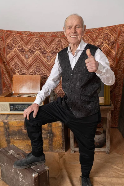 Счастливый улыбающийся старик в элегантной одежде, стоящий среди винтажной комнаты, кладет одну ногу на старый чемодан, держит большой палец вверх и смотрит в камеру — стоковое фото