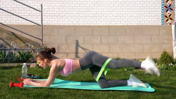 Mulher fitness fazendo exercício para glúteos com banda de resistência ao ar livre em um tapete de ioga. Treino de menina atlética — Vídeo de Stock