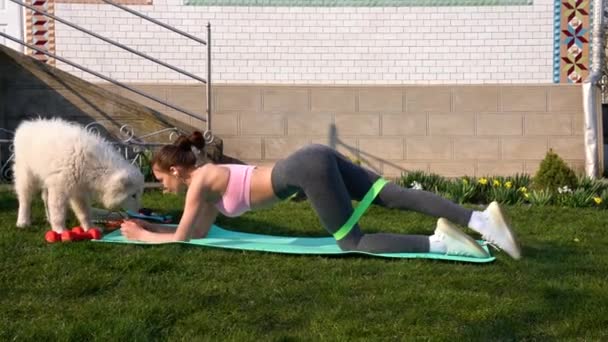 Trainieren Sie Frau mit Fitness-Elastikband-Workout im Freien zu Hause. Europäerin beim Widerstandstraining im Gras mit ihrem Samowar-Welpen. — Stockvideo