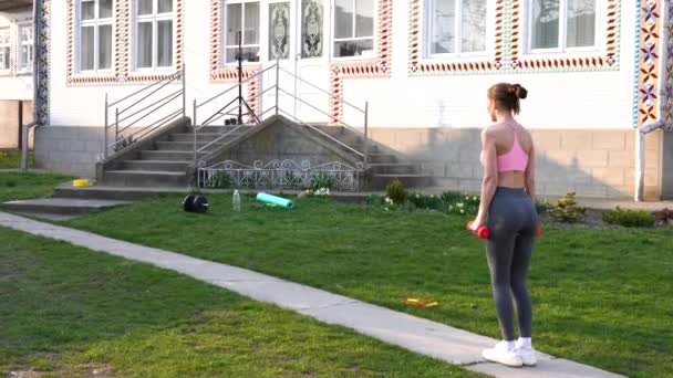 Jeune fille de fitness brune dans des vêtements de sport élégants faisant des fentes pour les jambes avec des haltères dans ses mains. Formation en plein air à domicile — Video