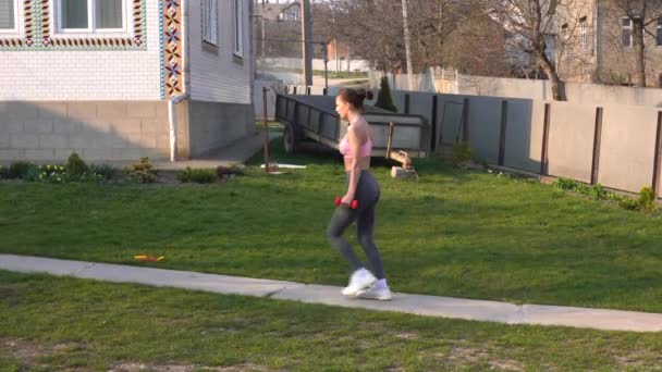 Vue latérale d'une jeune fille brune sportive en vêtements de sport faisant des exercices de squat avec des haltères rouges pour les fesses à l'extérieur à la maison . — Video