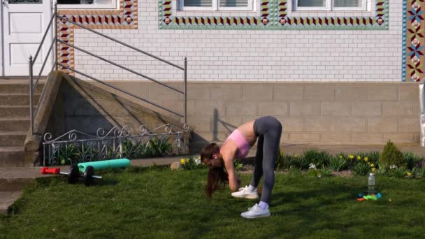 Schlankes brünettes Mädchen mit drahtlosen Kopfhörern in stylischer Sportbekleidung, die sich vor dem Heimtraining im Freien entwurmt und ausstreckt — Stockvideo