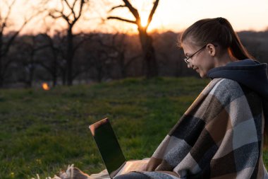 Parktaki sınıf projesi için çalışan pozitif üniversiteli kız. Gözlüklü güzel bir kadın çimenlerde oturuyor ve dizüstü bilgisayar kullanıyor..