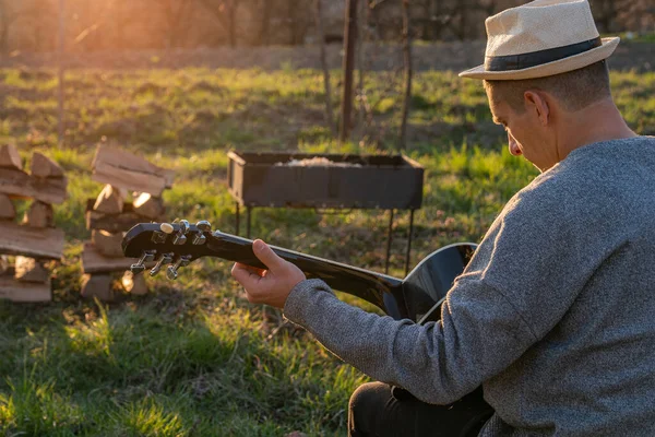 Mladý muž hraje na akustickou kytaru, zatímco sedí u táboráku. Dovolená koncept muž zpívá s kytarou na piknik — Stock fotografie