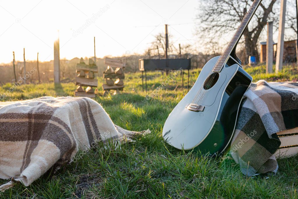 Black acoustic guitar on plaid, barbecue with firewood, sunset on the backgound