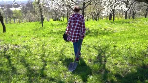 Уставшая женщина в клетчатой рубашке с лопатой и ведром в руках гуляет среди гаденов после тяжелого рабочего дня — стоковое видео